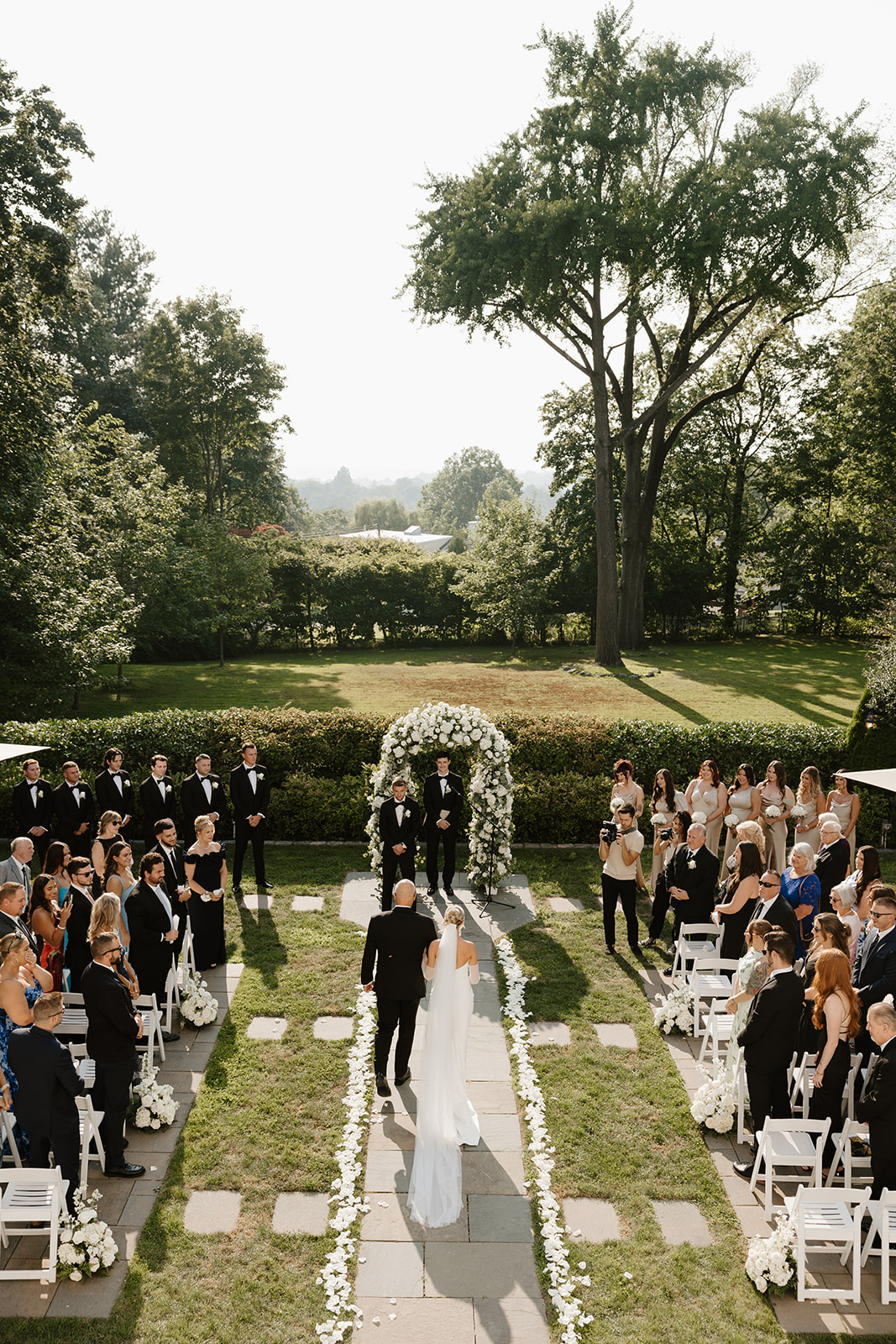 High-angle view of a bride walking down the stone aisle toward a white floral arch at an outdoor ceremony.